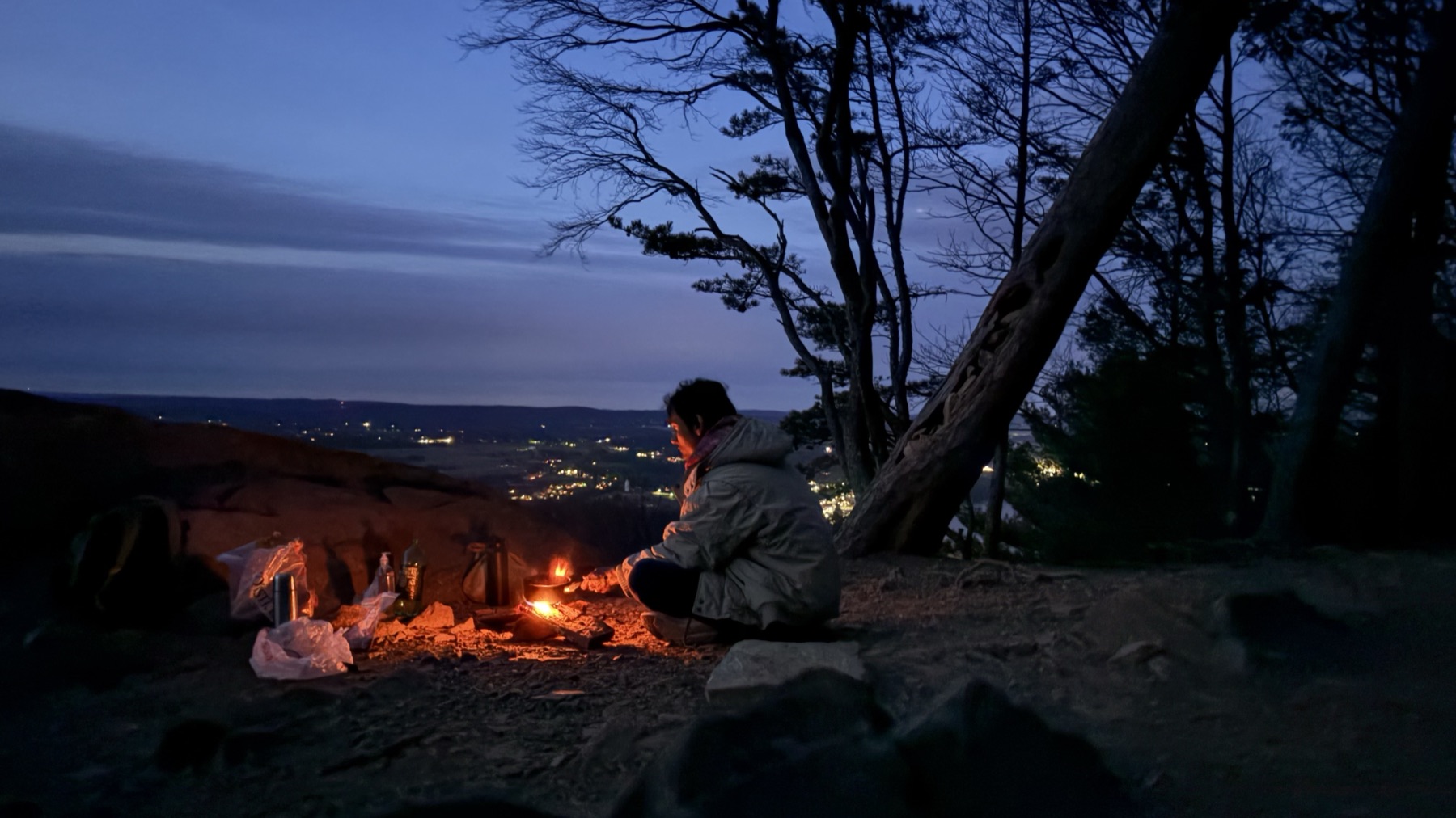 A small campfire on a mountain overlook at dusk, with town lights visible in the valley below